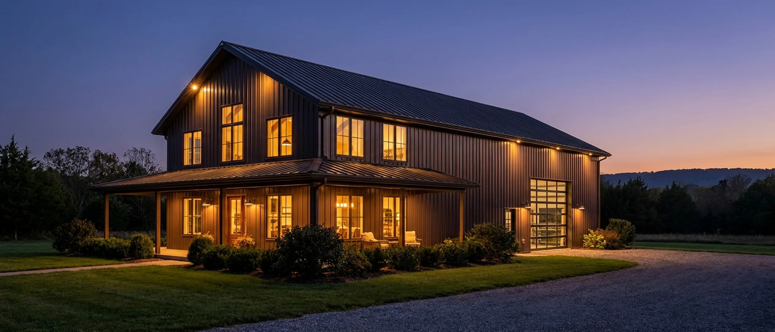 A barndominium at dusk with warm light glowing from every window and exterior lighting along the porch and roofline against a deep blue twilight sky.
